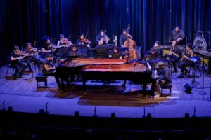 A young diverse music ensemble is on stage. There are two pianos at the center, facing each other so they are interlocking. Each is played by a young black man. Sheets of music have been tossed to the floor.