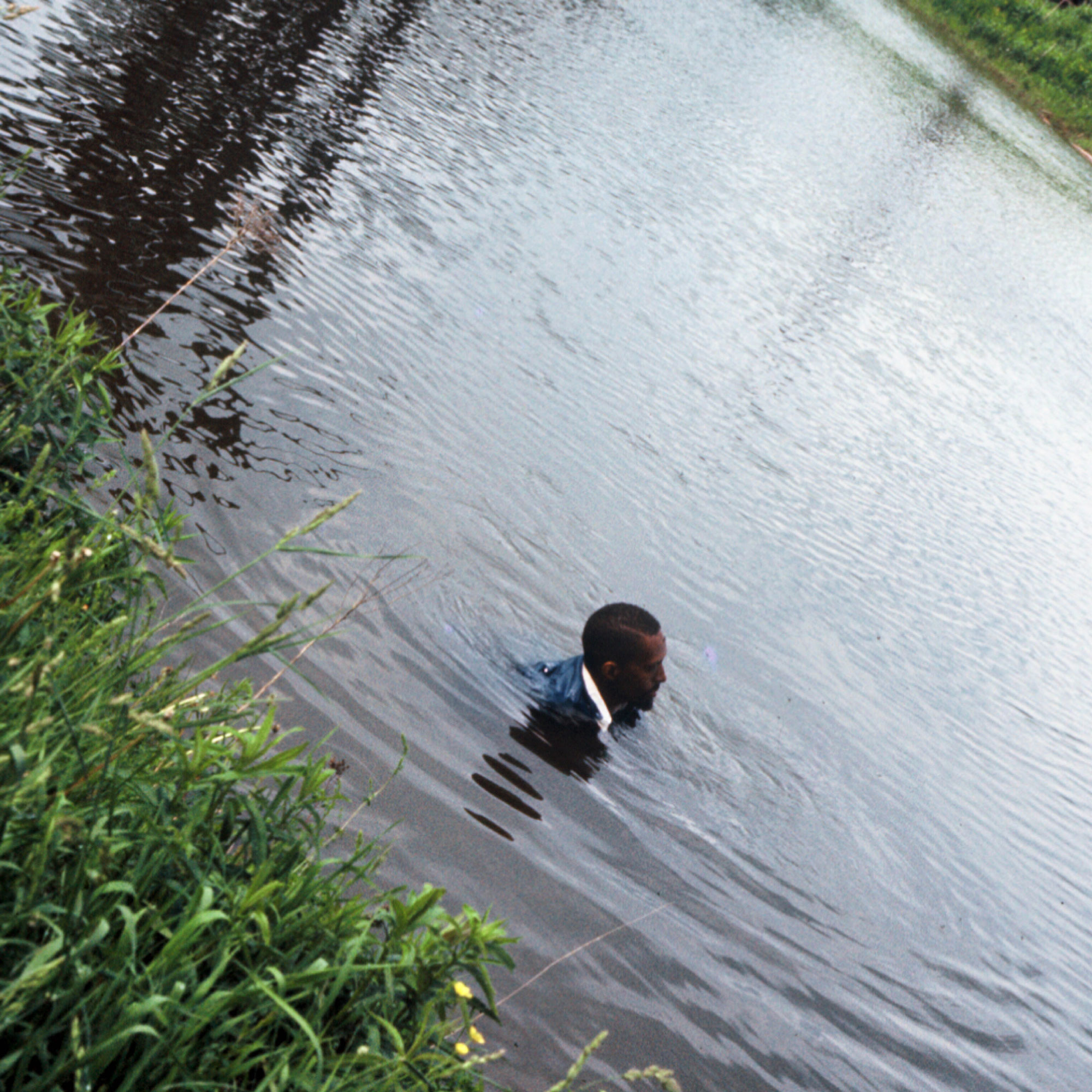 Julius Eastman, a black man, fully clothes and submerged in a lake with rings of movement in the water encircling his body